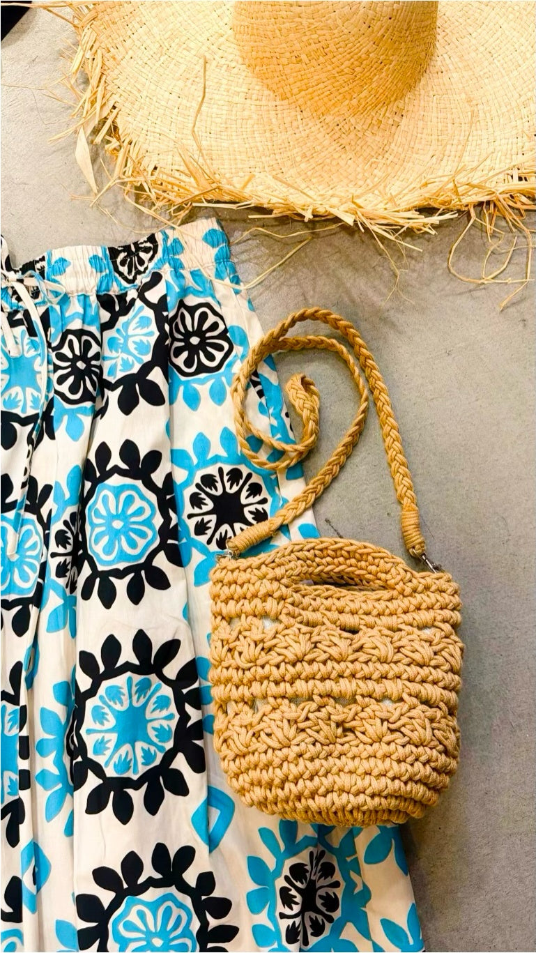 Dettaglio della gonna midi in popeline di cotone bianco con stampa a medaglioni floreali turchese e neri, vita elastica con coulisse, accostata a cappello di rafia e borsa crochet